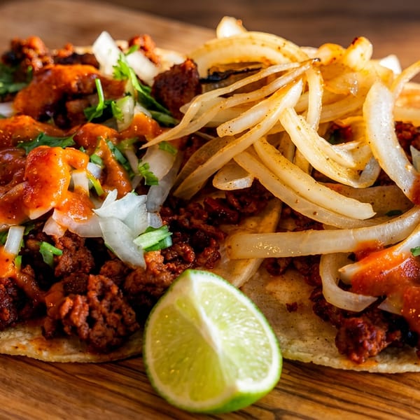 Two corn tortillas topped with seasoned meat, sautéed onions, cilantro, red sauce, and a lime wedge on a wooden table.